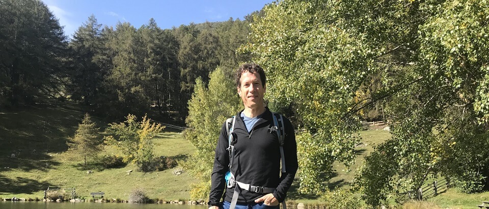 Ich bin mit meinem Wanderrucksack auf dem Rücken an einer Wiese mit Bäumen. Die Sonne scheint mir ins Gesicht.