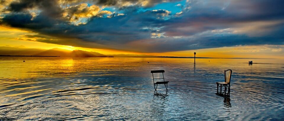 Nahe dem Ufer am Bodensee stehen zwei ungleiche Stühle im Wasser. Offenbar bedürfen die Stühle einer: Mediation.