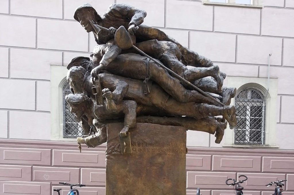 Bronzeskulptur von mehreren Menschen, die übereinander gestapelt sind. Text über Foto: „Nix gsagt ischt gnuag globat.“