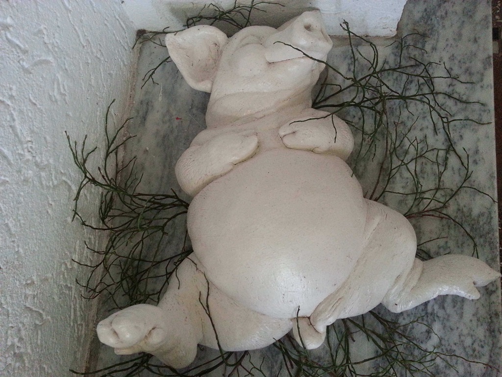 Weiße Skulptur eines liegenden Schweins mit geschlossenen Augen auf grünen dünnen Zweigen, Sinnbild für Entspannung.